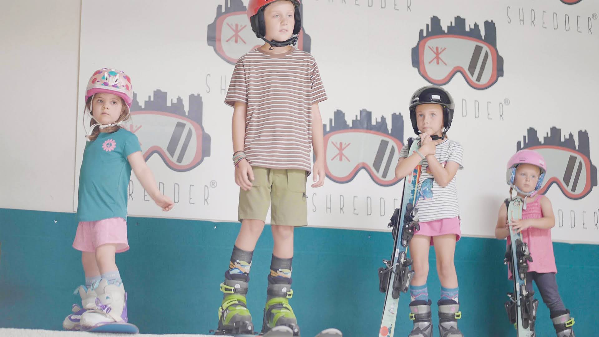 Group of kids in helmets standing in front of full Shredder logo wall, gear in hand