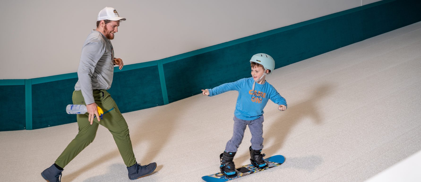 A coach walking alongside a young snowboarder on the indoor slope at Shredder North Side