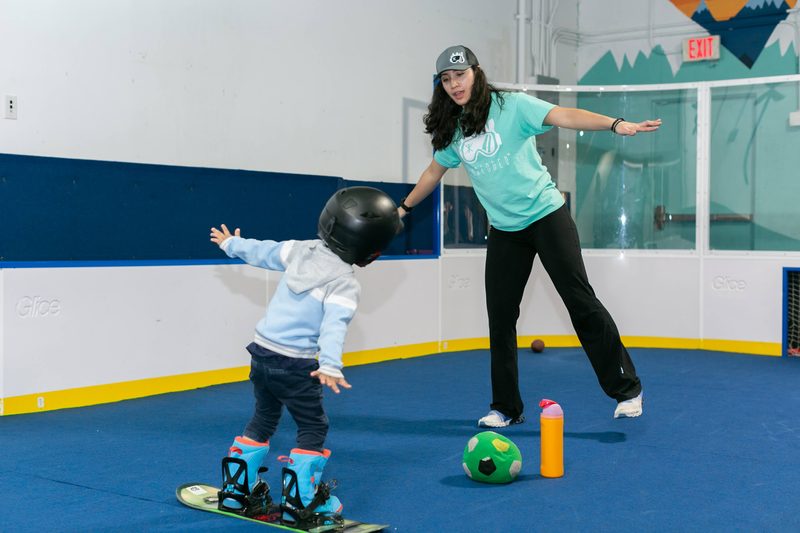 A coach demonstrating a stance while a toddler mirrors her on a snowboard