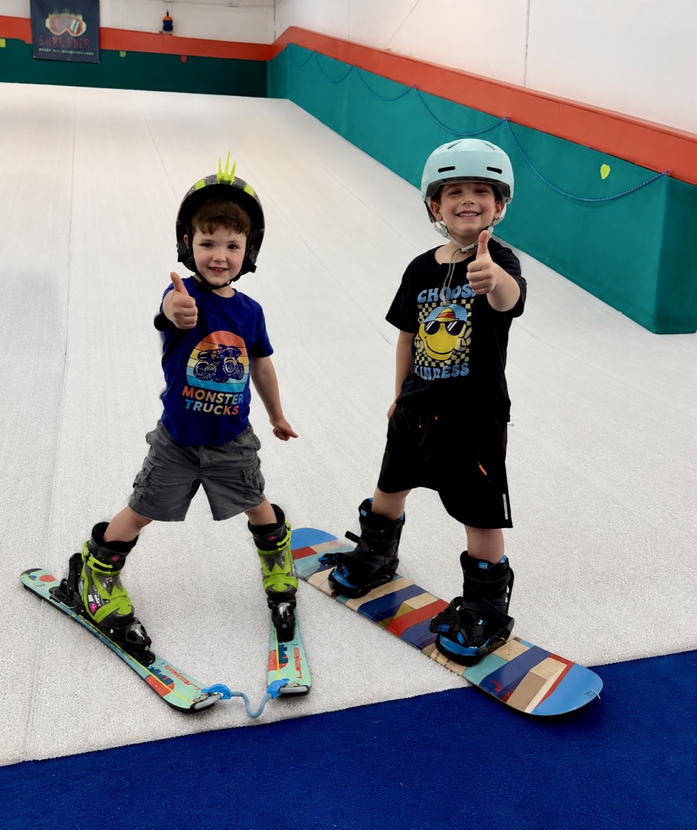 Two young Shredder students, one on skis and one on a snowboard, giving thumbs up on the indoor slope