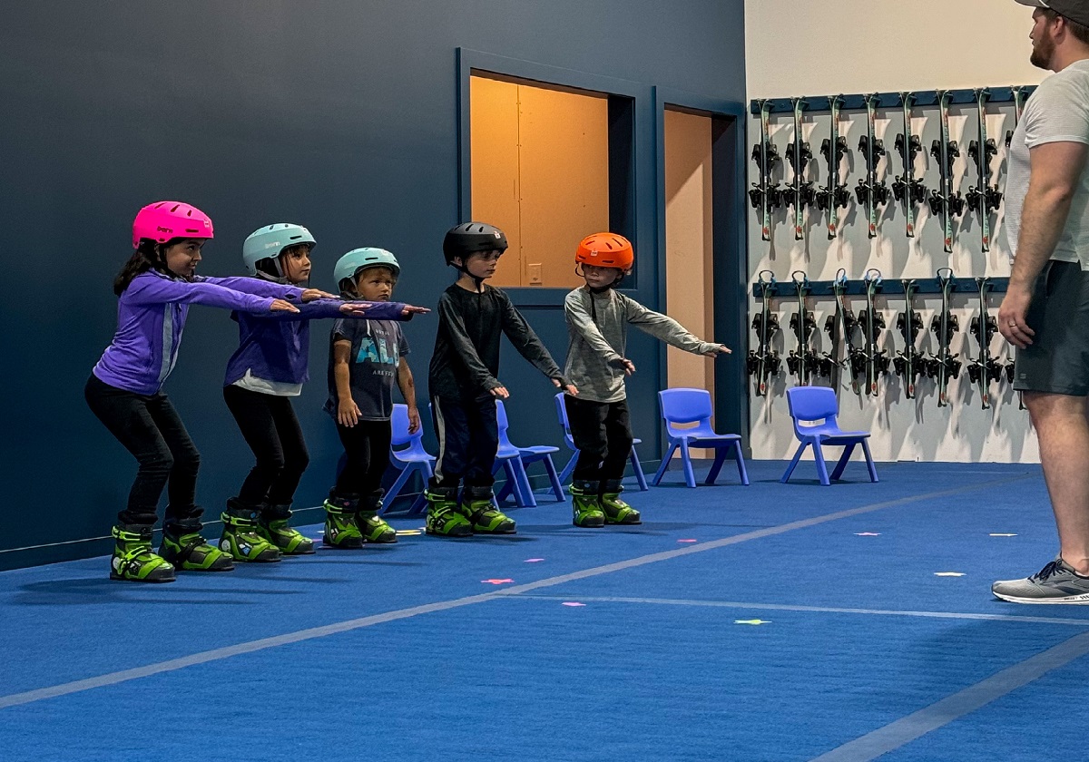 Kids in helmets learning the balance stance on the indoor slope with a coach