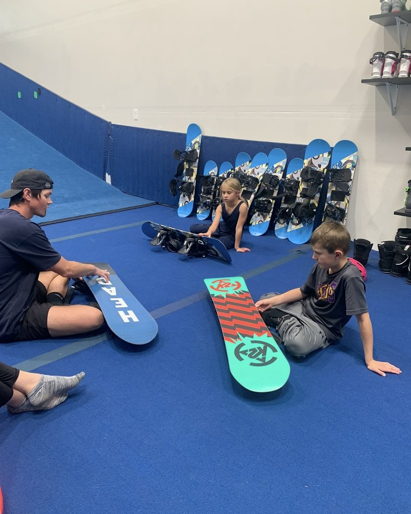 A coach sitting on the floor with two children at Shredder Lafayette, each with their own snowboard laid out in front of them