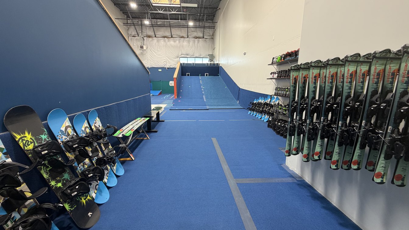 Children's snowboards racked along one wall and skis along the other, with the indoor slope between them at Shredder Lafayette