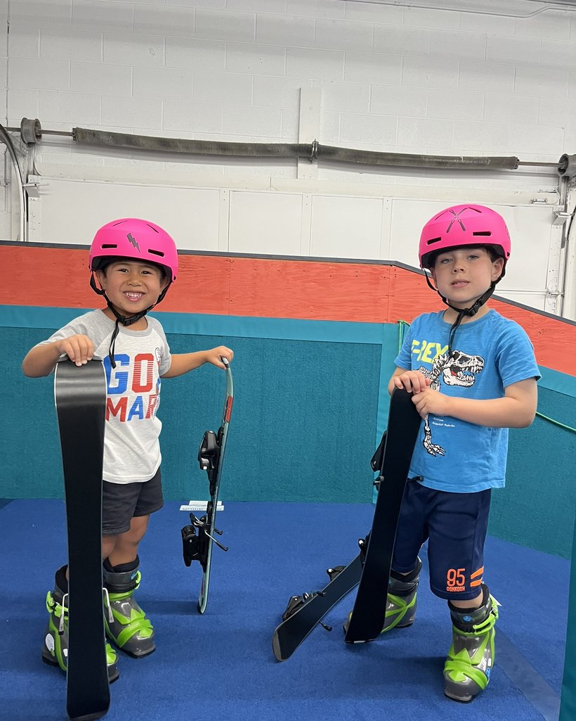 Two boys in fitted boots, each holding up their own snowboard and skis at Shredder Livonia