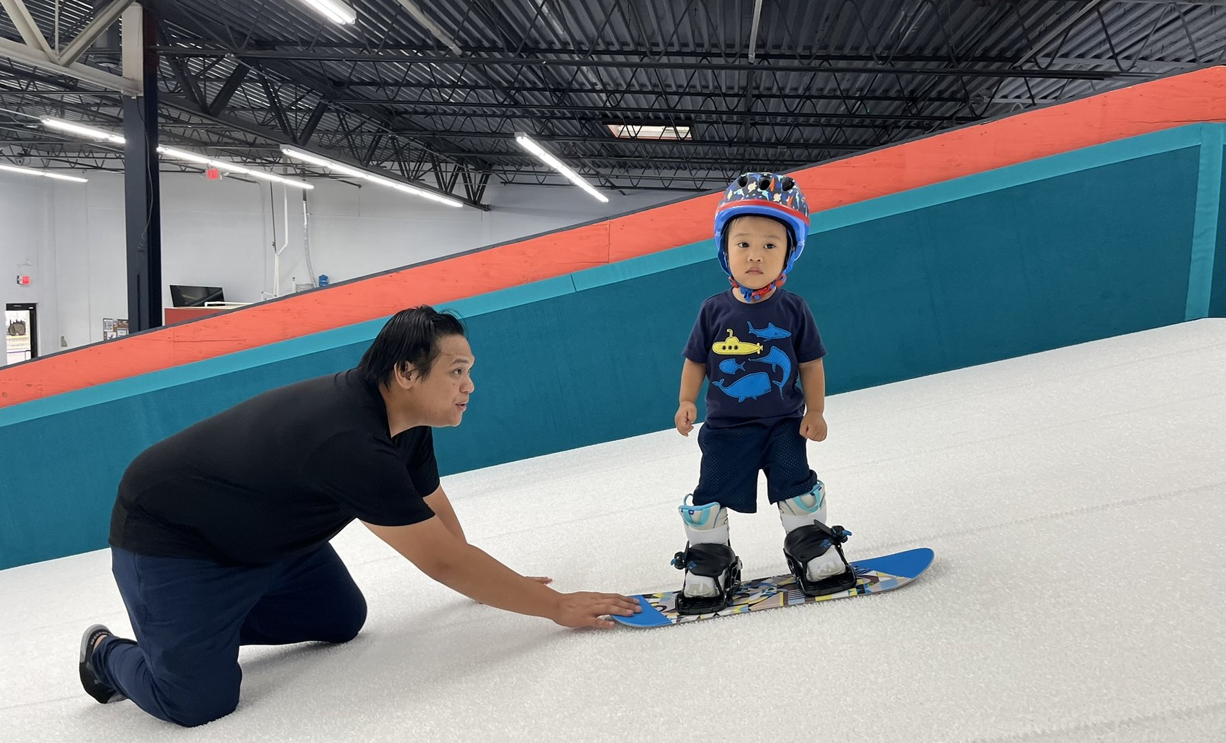 A coach kneeling on the indoor slope steadying a snowboard while a small child stands on it at Shredder Livonia