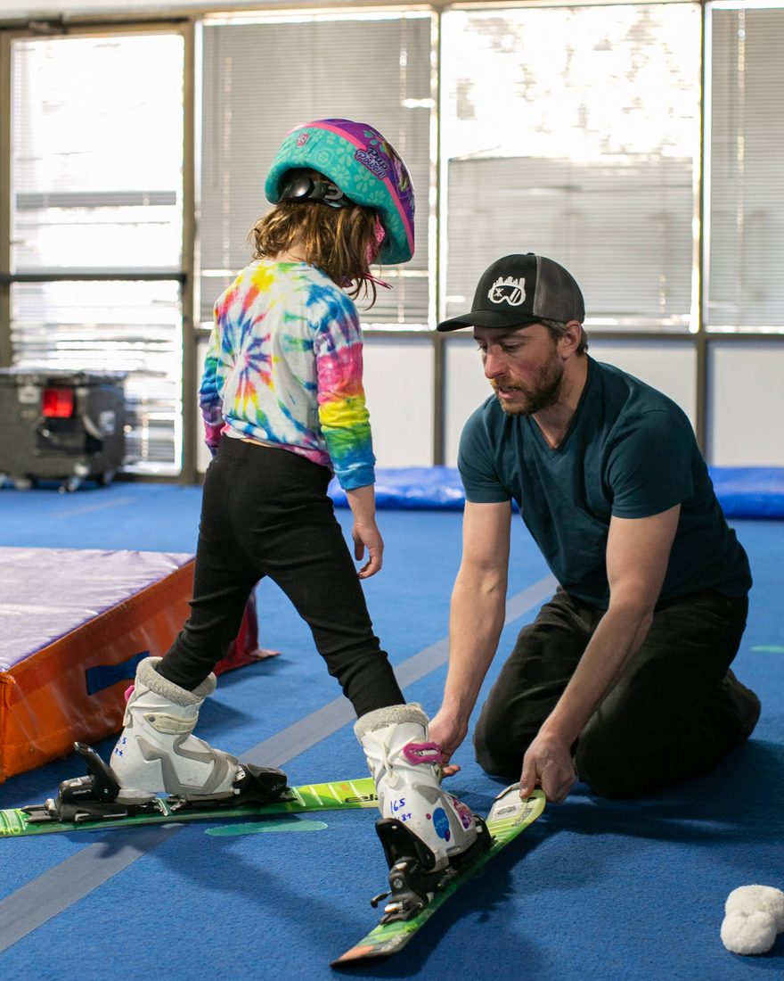 Illustration: a Shredder coach walking a parent and child through a rental setup — boots, helmet, goggles and gloves laid out on the bench.