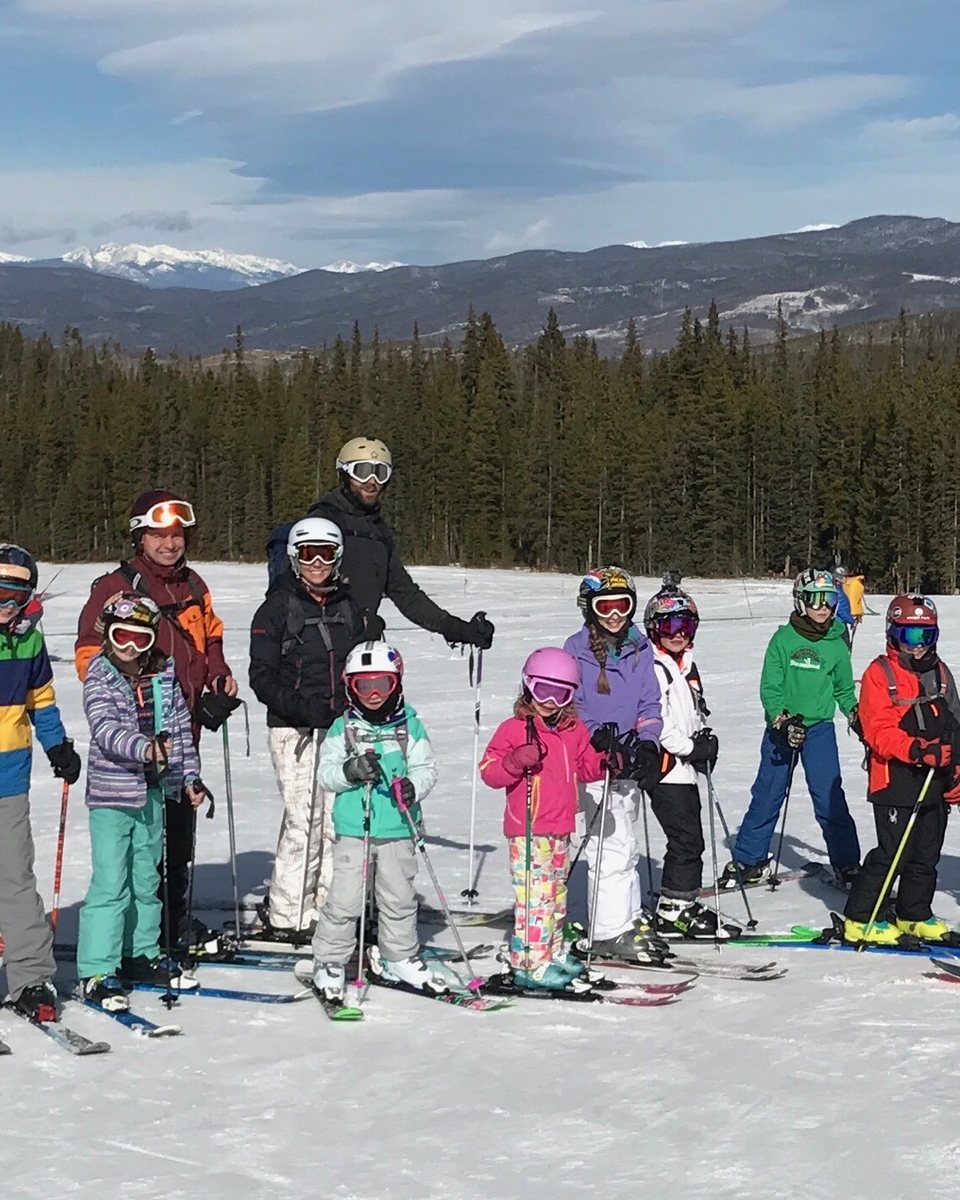 Shredder families together at the top of a run, skis on, the Rockies behind them