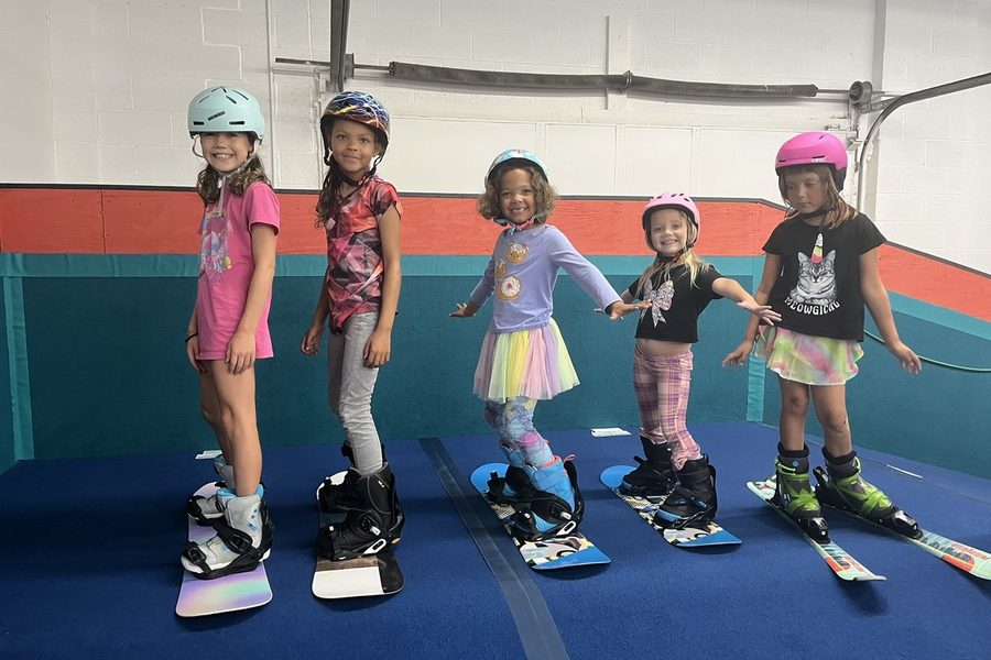 Five young kids standing in a row on the flat, boots strapped into snowboards and skis