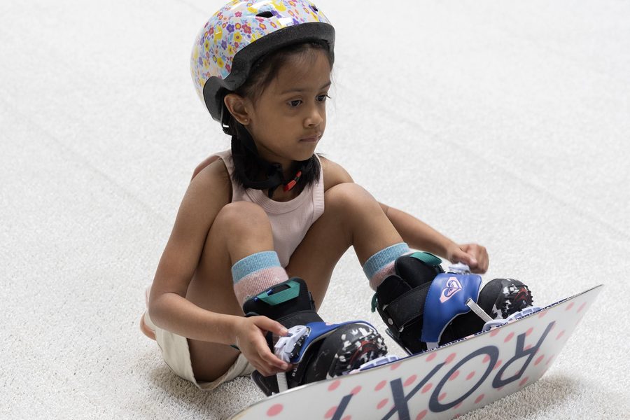 A child sitting on the snow strapping into a snowboard