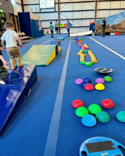 Two toddlers climbing soft mats beside a trail of colorful balance pods on a blue play floor