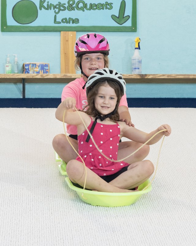A smiling young girl in a helmet sitting in a green sled, with an older girl behind her