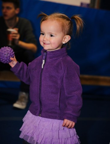A toddler in a purple jacket holding a soft spiky ball on the play floor at Shredder