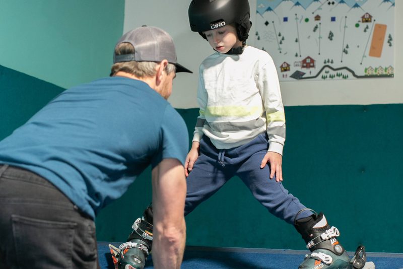 A coach guiding a young skier through a wedge stance on the indoor slope