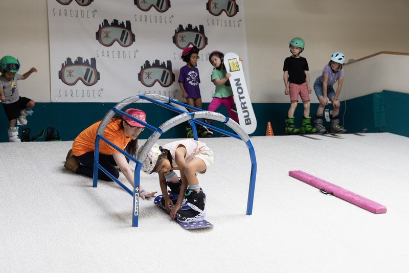 A coach kneeling with one young rider while three others wait their turn on the slope